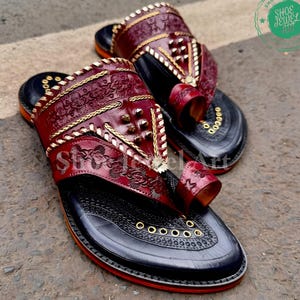 Puede incluir: Un par de sandalias de cuero hechas a mano con intrincados patrones florales y geom&eacute;tricos. Las sandalias presentan una parte superior de color burdeos oscuro con costuras y tachuelas doradas, una suela negra y una presilla para el dedo gordo. Las suelas tienen un ribete rojo. Las sandalias est&aacute;n etiquetadas como "Shoe Jewel Art".