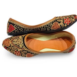 Könnte beinhalten: Ein Paar schwarze, bestickte Ballerinas mit Blumen- und Herzmotiven in Rot und Gold. Die Schuhe haben eine braune Ledereinfassung und eine beige Innenseite. Die Ballerinas sind wahrscheinlich für den Freizeitgebrauch.