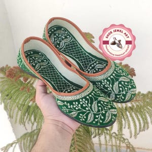 K&ouml;nnte beinhalten: Ein Paar gr&uuml;ne, bestickte Ballerinas mit floralem Design. Die Schuhe haben eine braune Einfassung und ein wei&szlig;es Innenfutter. Das Bild enth&auml;lt ein rosa-wei&szlig;es Logo mit der Aufschrift "Shoe Jewel Art Handmade Premium Shoes".