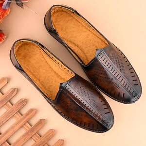 May include: A pair of brown leather slip-on shoes with intricate cut-out designs on the vamp. The shoes have a light brown interior and a dark brown exterior with a glossy finish. The shoes are displayed on a light brown surface.