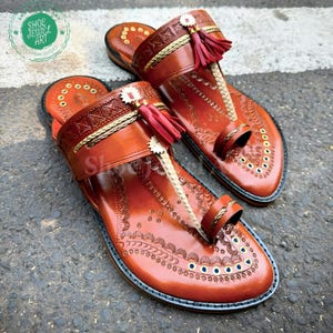 May include: A pair of handcrafted brown leather sandals with intricate embossed designs. The sandals feature a toe loop, decorative tassels, and gold and red accents. The sandals are from Shoe Jewel Art.