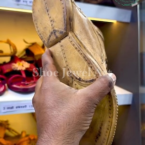 May include: Close-up of a tan leather shoe with intricate stitching and a textured sole. The shoe is held up, showcasing its design and craftsmanship. The image also shows other footwear on a shelf.