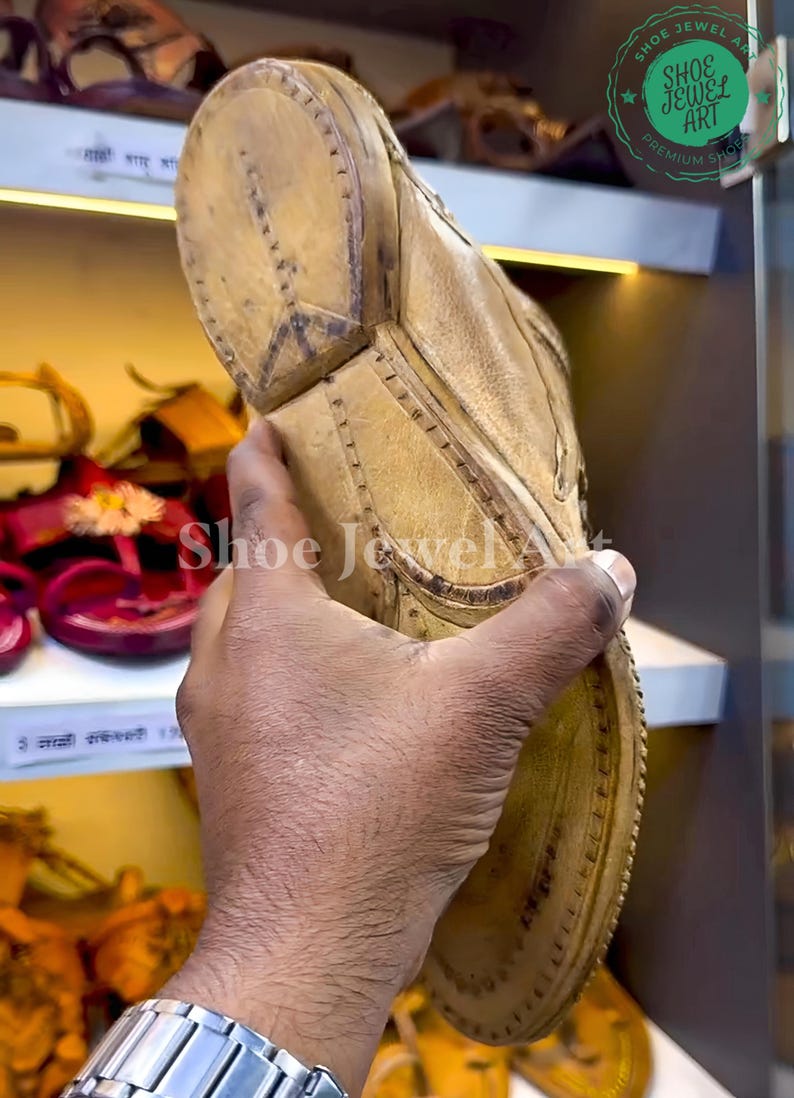May include: Close-up of a tan leather shoe with intricate stitching and a textured sole. The shoe is held up, showcasing its design and craftsmanship. The image also shows other footwear on a shelf.