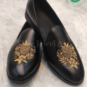 Puede incluir: Un par de mocasines de cuero negro con un diseño de escudo bordado en dorado en la pala. Los zapatos tienen una puntera redondeada y un tacón bajo. La imagen también incluye un logotipo que dice "Shoe Jewel Art" y "Premium Shoes".