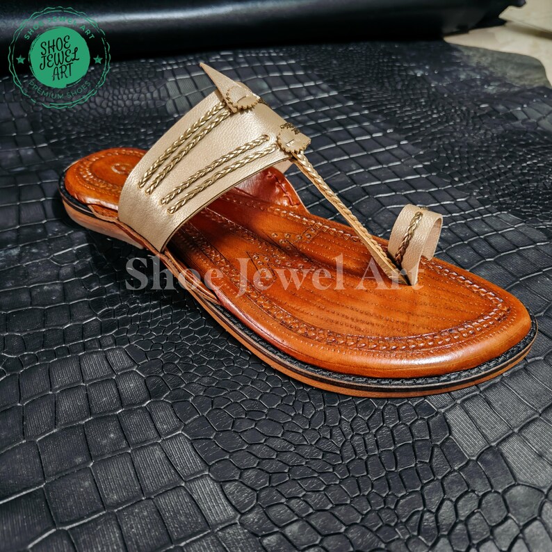 May include: A handcrafted leather sandal with a light brown sole and a beige upper adorned with gold-colored braided accents. The sandal features a toe loop and a strap design. The words "Shoe Jewel Art" are visible.