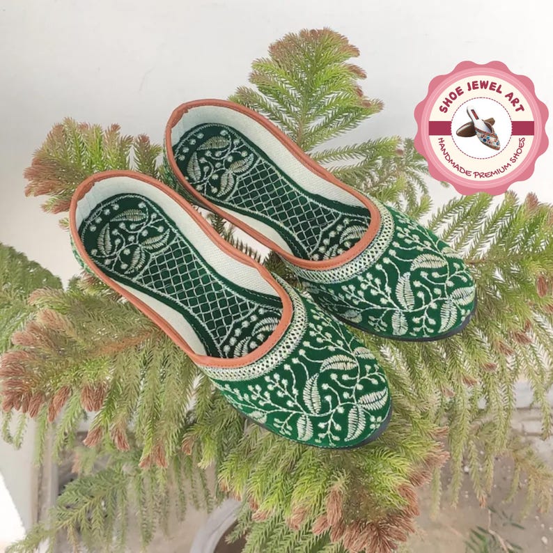 K&ouml;nnte beinhalten: Ein Paar gr&uuml;ne, bestickte Ballerinas mit wei&szlig;en Blumenmustern und einem beigen Besatz. Die Schuhe werden auf einem Bett aus gr&uuml;nem und braunem Laubwerk pr&auml;sentiert. Das Bild enth&auml;lt ein rosa-wei&szlig;es Logo mit der Aufschrift "Shoe Jewel Art, Handmade Premium Shoes."