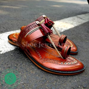 May include: A pair of brown leather sandals with intricate embossed designs. The sandals feature a toe loop and a strap with a decorative tassel. The text "Shoe Jewel Art" is visible on the sandals.