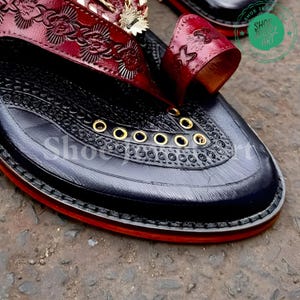 Puede incluir: Primer plano de un zapato de cuero negro con intrincados patrones en relieve y un ribete rojo. El zapato presenta una correa de cuero burdeos con dise&ntilde;os florales y de estrellas, y ojales dorados. El texto "Shoe Art" es visible.