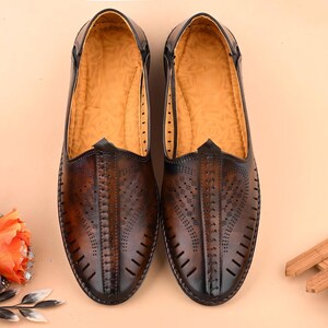 May include: A pair of brown leather slip-on shoes with intricate cut-out designs on the vamp. The shoes have a pointed toe and a dark brown trim. The interior is a light tan color. The shoes are displayed on a light tan surface.