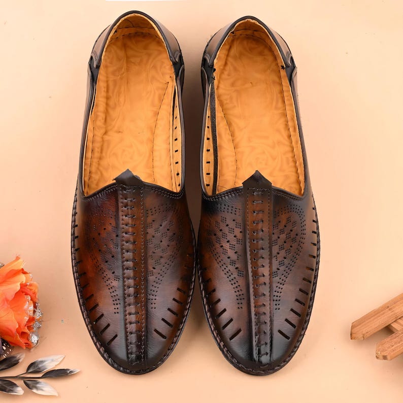 May include: A pair of brown leather slip-on shoes with intricate cut-out designs on the vamp. The shoes have a pointed toe and a dark brown trim. The interior is a light tan color. The shoes are displayed on a light tan surface.