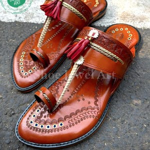 May include: A pair of brown leather sandals with intricate embossed patterns and red tassel accents. The sandals feature a toe loop and a strap across the top of the foot. The soles are outlined in black. The sandals are labeled "Shoe Jewel Art Premium Shoes".