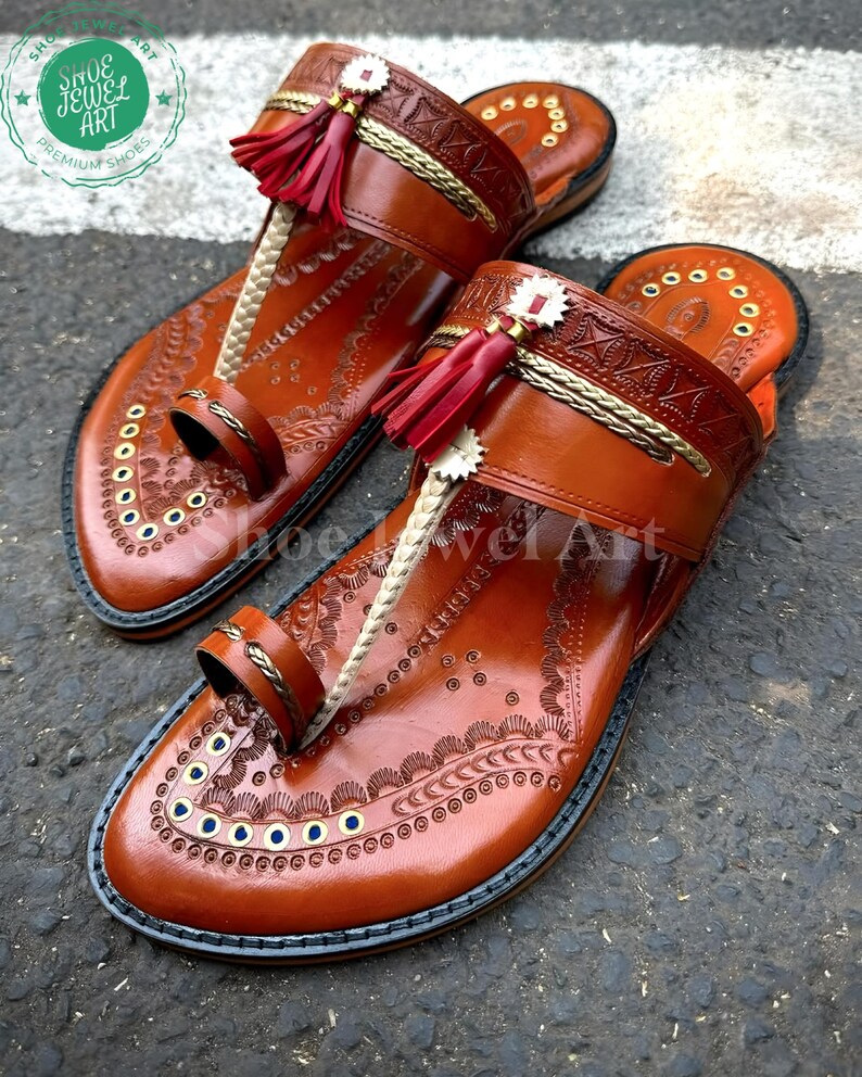 May include: A pair of brown leather sandals with intricate embossed patterns and red tassel accents. The sandals feature a toe loop and a strap across the top of the foot. The soles are outlined in black. The sandals are labeled "Shoe Jewel Art Premium Shoes".