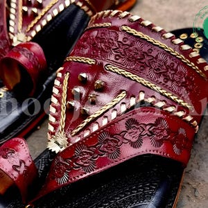 Puede incluir: Primer plano de un par de sandalias de cuero hechas a mano. Las sandalias presentan una parte superior de color rojo oscuro con intrincados patrones florales en relieve, detalles dorados y tachuelas met&aacute;licas. Las suelas negras tienen detalles dorados, mostrando una mezcla de artesan&iacute;a tradicional y dise&ntilde;o moderno.