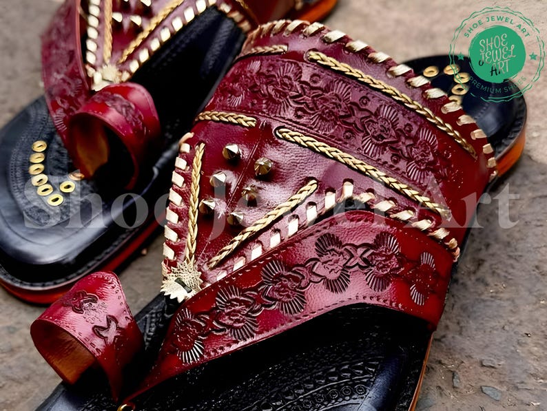 Puede incluir: Primer plano de un par de sandalias de cuero hechas a mano. Las sandalias presentan una parte superior de color rojo oscuro con intrincados patrones florales en relieve, detalles dorados y tachuelas met&aacute;licas. Las suelas negras tienen detalles dorados, mostrando una mezcla de artesan&iacute;a tradicional y dise&ntilde;o moderno.