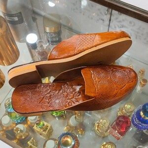 May include: A pair of brown leather slip-on sandals. The sandals feature a floral embossed design on the footbed and a textured pattern on the top strap. The soles are a light brown color.