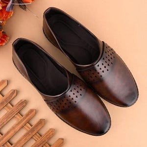 Op de afbeelding: Een paar bruine leren instappers. De schoenen hebben een ronde neus en een decoratief ruitvormig uitgesneden patroon aan de bovenkant. De schoenen worden gepresenteerd op een lichtbruin oppervlak.
