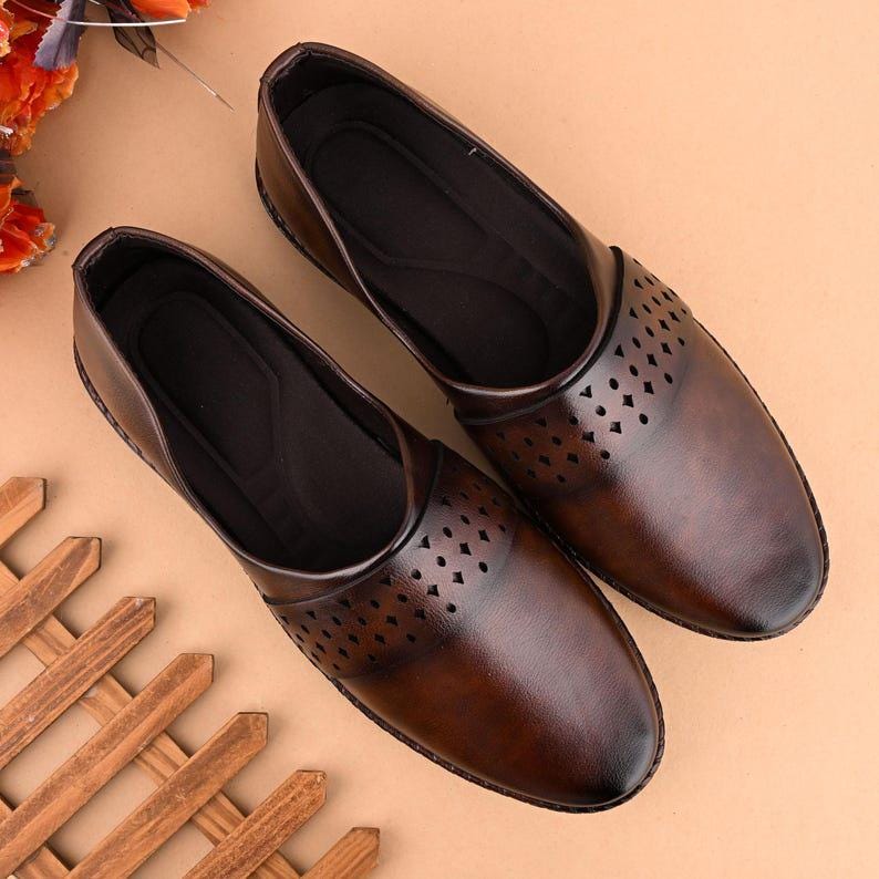 Op de afbeelding: Een paar bruine leren instappers. De schoenen hebben een ronde neus en een decoratief ruitvormig uitgesneden patroon aan de bovenkant. De schoenen worden gepresenteerd op een lichtbruin oppervlak.