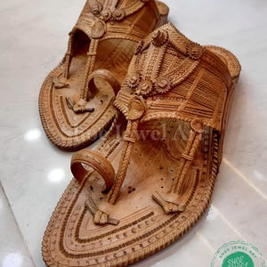 May include: A pair of intricately carved, light brown leather sandals. The sandals feature detailed patterns, including floral and geometric designs, with a toe loop and ankle strap. The soles are also decorated with carved details. The sandals are displayed on a light-colored surface.