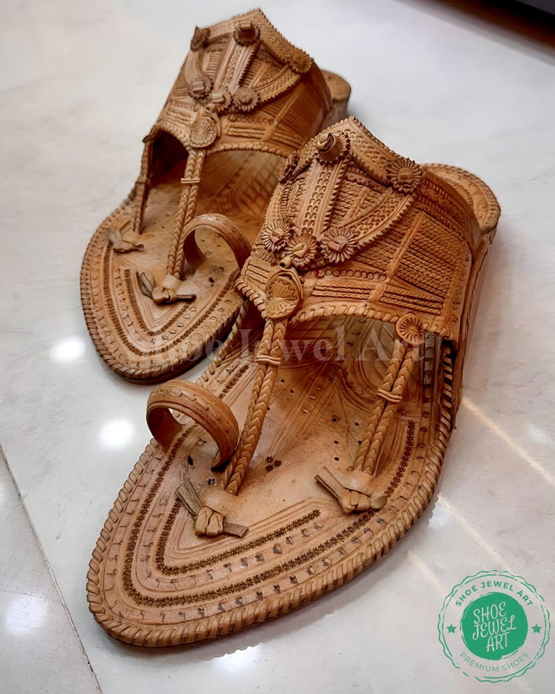 May include: A pair of intricately carved, light brown leather sandals. The sandals feature detailed patterns, including floral and geometric designs, with a toe loop and ankle strap. The soles are also decorated with carved details. The sandals are displayed on a light-colored surface.