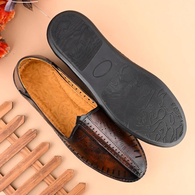 May include: A pair of brown leather loafers with a textured design. One shoe is right-side up, displaying the perforated detailing and stitching. The other shoe is upside down, revealing the black sole. The shoes are on a light brown surface.
