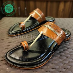 May include: Pair of black leather sandals with brown leather straps and gold-colored accents. The sandals feature a toe loop and a decorative strap across the top of the foot. The soles are black with a brown border. The sandals are labeled "Shoe Jewel Art".