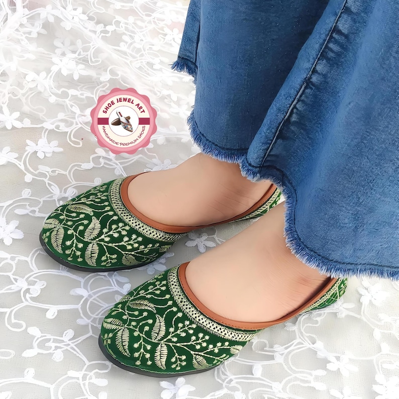 K&ouml;nnte beinhalten: Gr&uuml;ne, bestickte Slipper mit Blattmuster und braunem Besatz. Die Schuhe werden mit blauen Jeans getragen. Die Schuhe haben eine runde Spitze und eine flache Sohle. Die Schuhe sind handgefertigt.