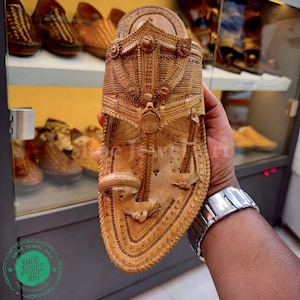 May include: A handcrafted, light brown leather sandal with intricate carved details. The sandal features floral and geometric patterns, with a decorative top piece. The sole is also detailed with a stitched border. The sandal is displayed in a shop setting.