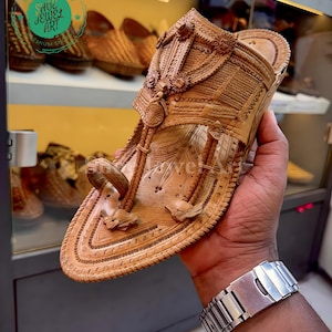 May include: A handcrafted, light brown leather sandal with intricate carved details. The sandal features a toe loop and decorative elements. The image also shows other similar sandals in the background.