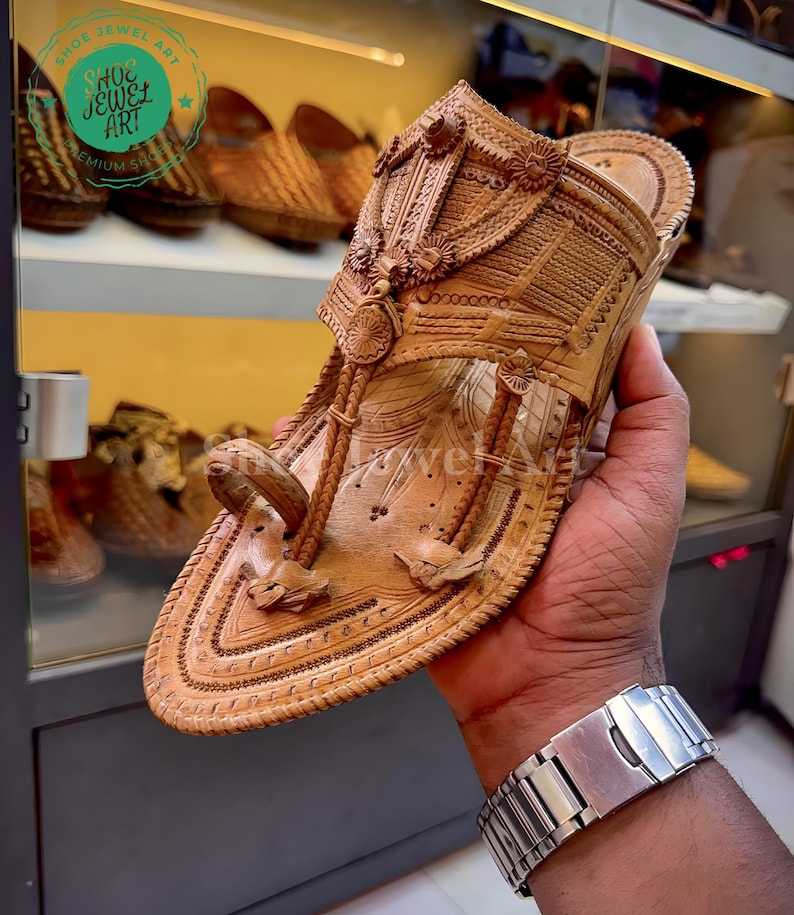 May include: A handcrafted, light brown leather sandal with intricate carved details. The sandal features a toe loop and decorative elements. The image also shows other similar sandals in the background.