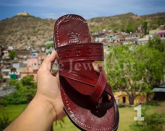 May include: A burgundy leather sandal with a stitched sole and decorative cross straps. The sandal features a textured surface and a dark brown border. The background shows a green landscape and a blue sky.