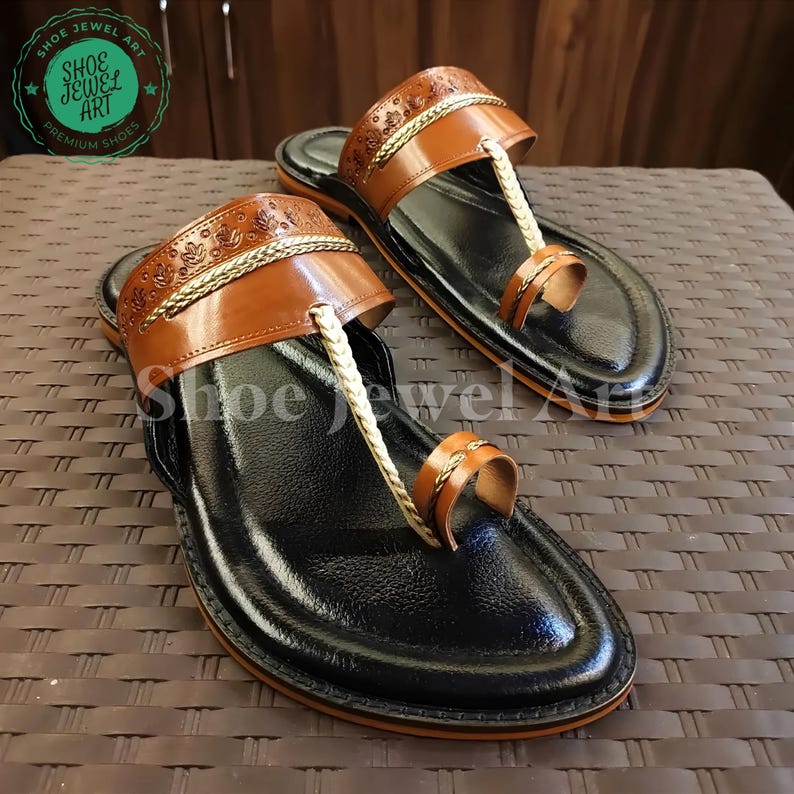 May include: A pair of black leather sandals with brown leather straps and gold-colored accents. The sandals have a toe loop and a strap across the top of the foot. The brand name "Shoe Jewel Art" is visible in a circular logo.