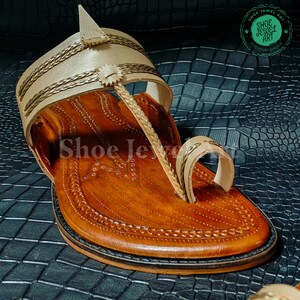 May include: Handmade leather sandal with intricate stitching and braided accents. The sandal features a light tan upper with gold-colored trim and a brown sole. The text "Shoe Jewel Art" is visible on the sandal.