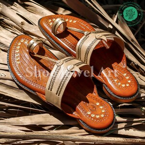 May include: A pair of brown leather sandals with beige straps and decorative stitching. The sandals feature a toe loop and a strap across the top of the foot. The soles are dark brown, and the sandals are decorated with small gold-colored rivets. The sandals are displayed on a bed of dried leaves.