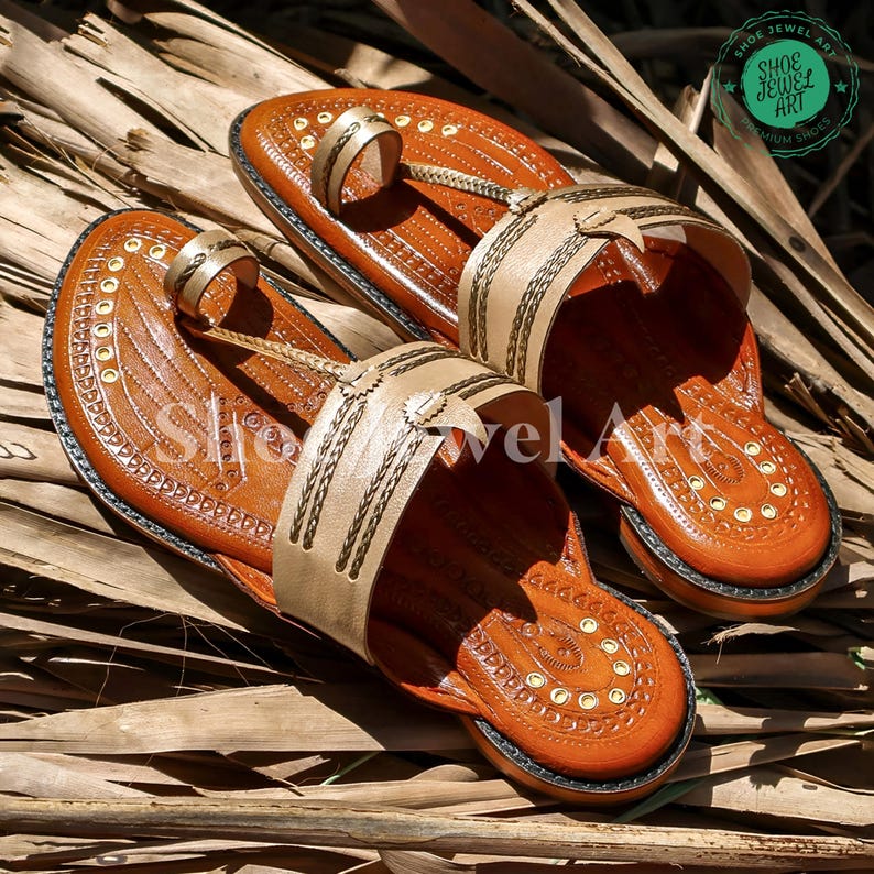 May include: A pair of brown leather sandals with beige straps and decorative stitching. The sandals feature a toe loop and a strap across the top of the foot. The soles are dark brown, and the sandals are decorated with small gold-colored rivets. The sandals are displayed on a bed of dried leaves.