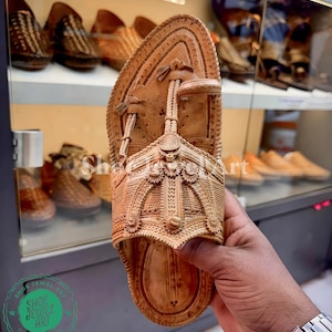 May include: A light brown leather sandal with intricate carved details. The sandal is held up, showcasing the detailed design and craftsmanship. Other similar sandals are visible in the background.
