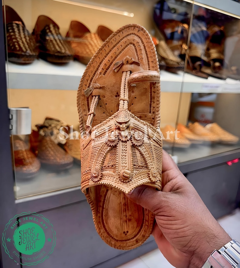 May include: A light brown leather sandal with intricate carved details. The sandal is held up, showcasing the detailed design and craftsmanship. Other similar sandals are visible in the background.