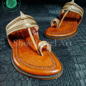 May include: A pair of handcrafted leather sandals with a light tan upper and a brown footbed. The sandals feature intricate stitching and a toe loop design. The soles are dark brown with a black border. The sandals are displayed on a textured black surface.