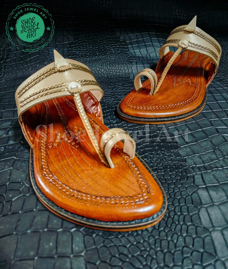May include: A pair of handcrafted leather sandals with a light tan upper and a brown footbed. The sandals feature intricate stitching and a toe loop design. The soles are dark brown with a black border. The sandals are displayed on a textured black surface.