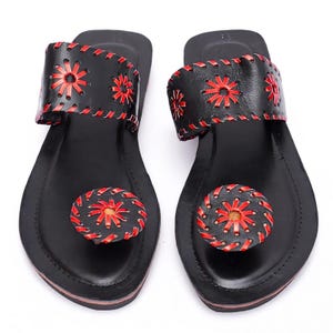 May include: A pair of black leather sandals with red floral accents. The sandals feature a thong design with a strap across the top of the foot and a circular design on the toe. The edges are stitched with red thread.