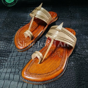 May include: A pair of handcrafted leather sandals with a tan upper and a brown sole. The sandals feature intricate stitching and a toe loop design. The upper has gold-colored accents and a decorative strap. The sandals are displayed on a textured black surface.