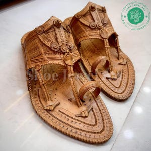May include: A pair of intricately carved, light brown leather sandals. The sandals feature detailed floral and geometric patterns, with a toe loop and ankle strap design. The image includes the text "SHOE JEWEL ART PREMIUM SHOES".