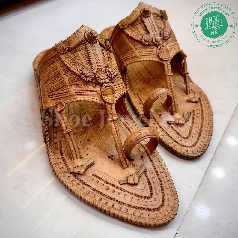 May include: A pair of intricately carved, light brown leather sandals. The sandals feature detailed floral and geometric patterns, with a toe loop and ankle strap design. The image includes the text "SHOE JEWEL ART PREMIUM SHOES".