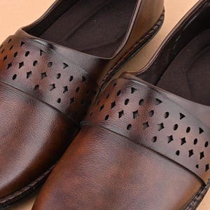 Op de afbeelding: Close-up van twee bruine leren instappers. De schoenen hebben een decoratieve band over de bovenkant met verschillende uitgesneden vormen. Het leer heeft een gepolijste afwerking en de schoenen lijken ongedragen.