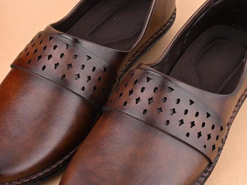 Op de afbeelding: Close-up van twee bruine leren instappers. De schoenen hebben een decoratieve band over de bovenkant met verschillende uitgesneden vormen. Het leer heeft een gepolijste afwerking en de schoenen lijken ongedragen.