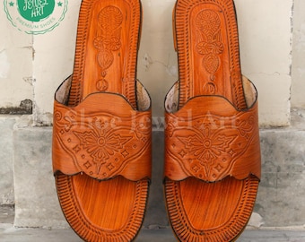 May include: A pair of handcrafted, open-toe leather sandals in a warm orange hue. The sandals feature intricate embossed floral and geometric designs on the straps and footbeds. The sandals are labeled "Shoe Jewel Art Premium Shoes".