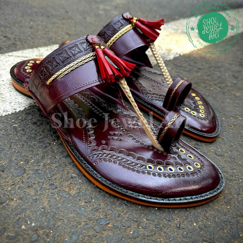 May include: Burgundy leather sandals with intricate embossed patterns and gold accents. The sandals feature a toe loop, decorative tassels, and the text "Shoe Jewel Art".