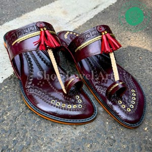 May include: A pair of burgundy leather sandals with intricate embossed patterns and gold accents. The sandals feature a toe loop, decorative tassels, and a gold trim. The soles have an orange border and the text "Show Jewel Art".