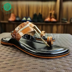 May include: Black leather sandals with brown leather straps and accents. The sandals have a toe loop and a decorative strap across the top of the foot. The soles have an orange trim. The sandals are displayed on a textured surface.