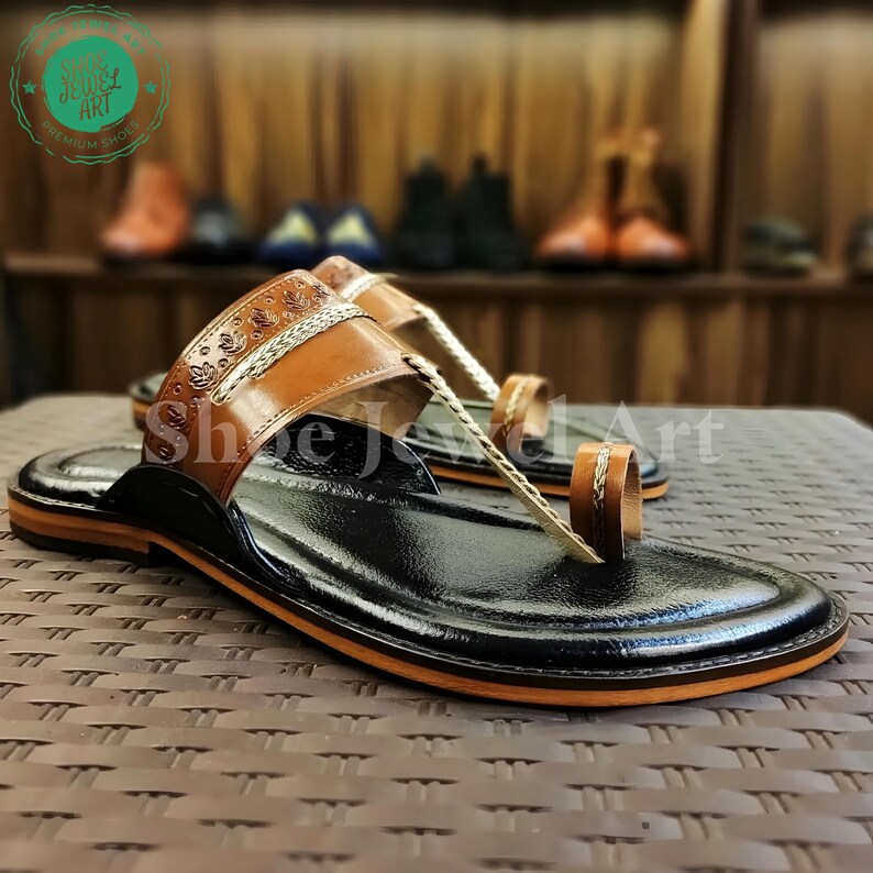 May include: Black leather sandals with brown leather straps and accents. The sandals have a toe loop and a decorative strap across the top of the foot. The soles have an orange trim. The sandals are displayed on a textured surface.