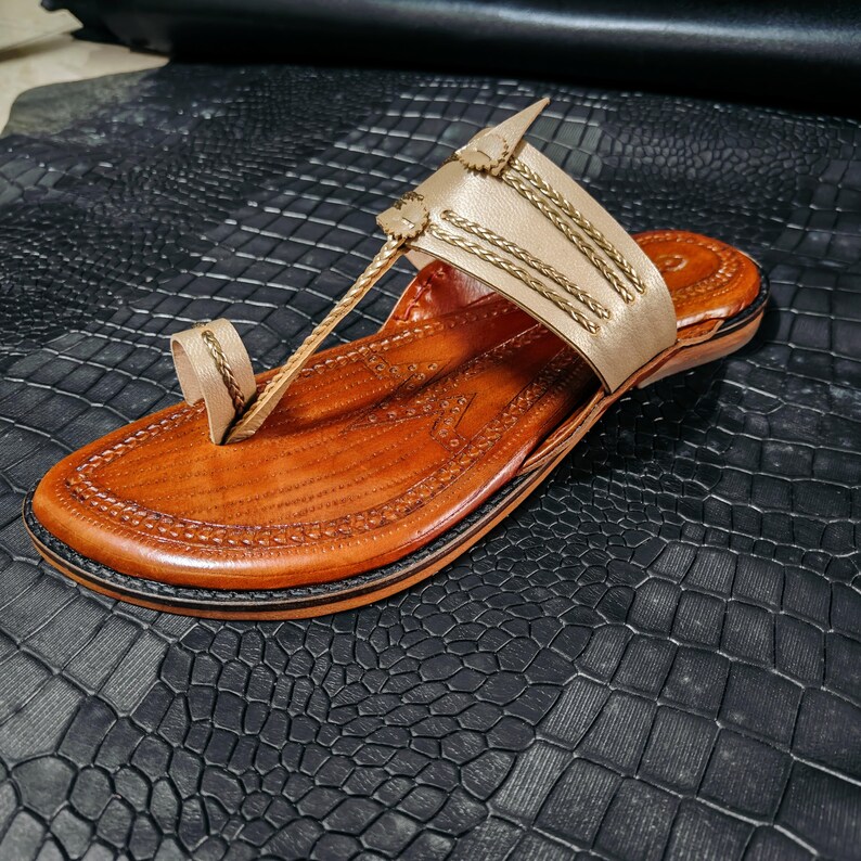 May include: A handcrafted leather sandal with a light brown upper and a darker brown sole. The sandal features decorative stitching and gold-colored accents. The toe strap and upper strap are connected by a thin leather strap.